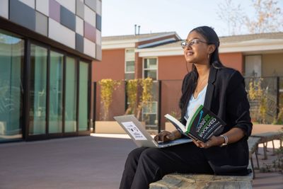 Student outside working on a laptop while holding a book.