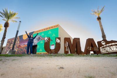 An N A U student makes a Y with her arms in the air next to a sign to spell out Yuma.